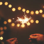 Happy New Year. Glowing sparklers in hands on background of golden christmas tree lights, couple celebrating in dark festive room. Space for text. Fireworks burning in hands. Happy Holidays